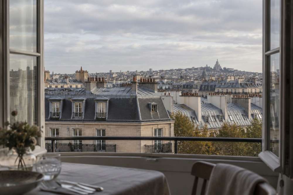 Toits de zinc et cheminées de Paris sous un ciel de fin de journée, vus par une grande fenêtre ouverte, avec une table dressée au premier plan.